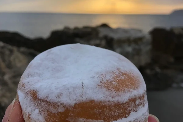 Krapfen spolverato di zucchero a velo tenuto in mano al tramonto sulla spiaggia.