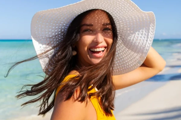 Donna sorridente con cappello bianco in spiaggia soleggiata.